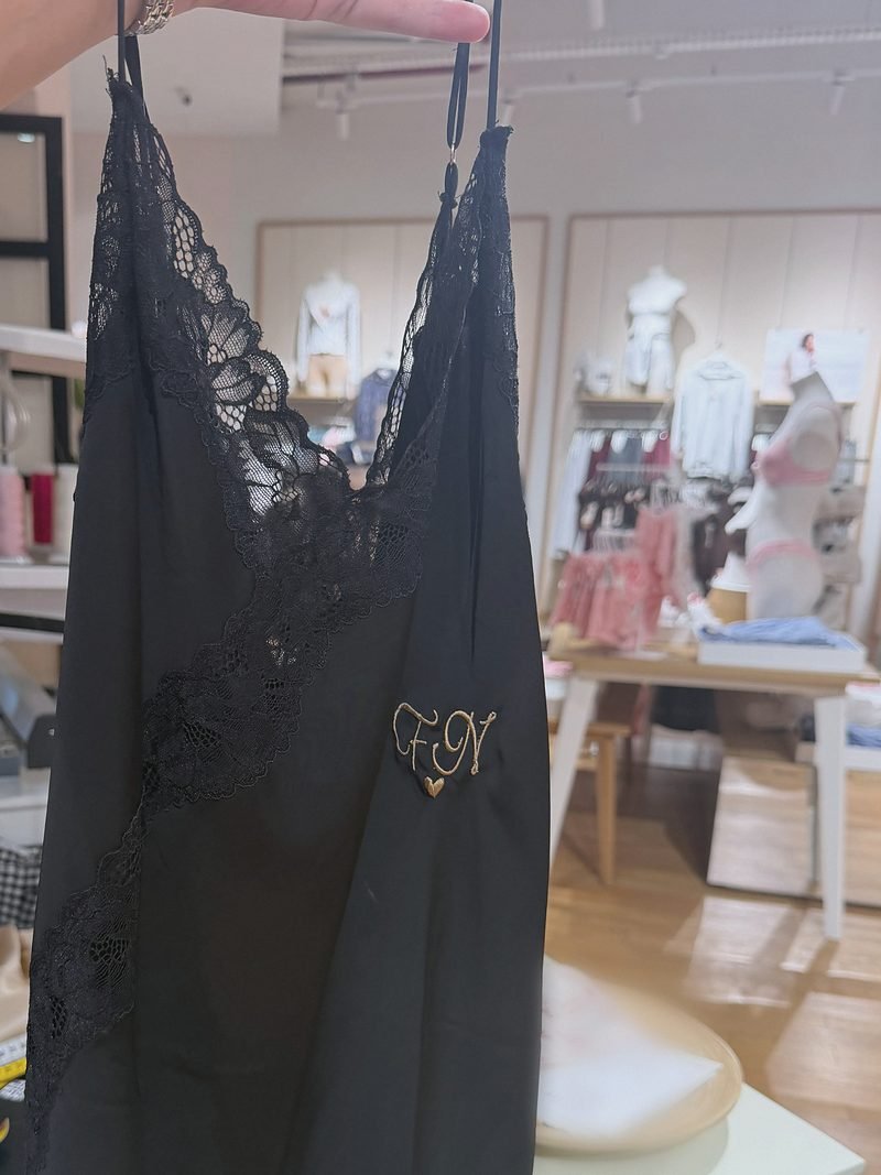 Nuisette noire en dentelle sur cintre, initiales brodées en fil doré, boutique en arrière-plan